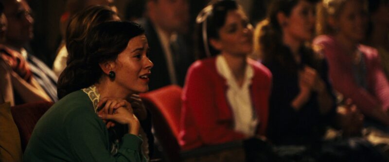 Women looking in awe. One in a green sweater. One in a red sweater. Other women surround them. 